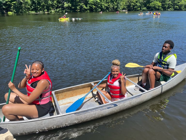 Three people in a rowboat on a river with green trees in the background. Two children and one adult hold paddles and wear life jackets.