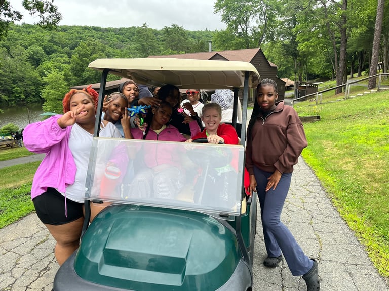 Jacqueline McDonough smiling with campers in a golf cart on a park path with green lawns and trees in the background