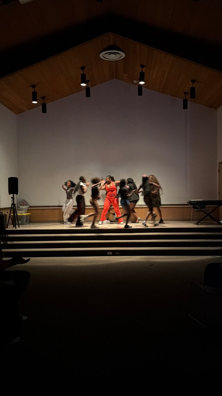 A group of dancers performing on stage under dramatic lighting, with one dancer in a red dress center stage surrounded by dancers in dark clothing