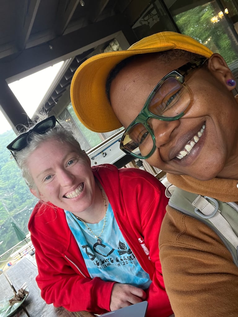 Two people smiling at camera inside a vehicle, one wearing a yellow cap and glasses, the other in a red jacket